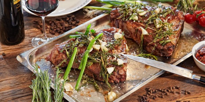 Steak auf Backblech. Andekoriert mit Rosmarin, Frühlingszwiebeln und Knoblach. Im Anschnitt liegt ein Messer welches mit der Messerspitze auf dem Fleisch liegr. Im Hintergrund sieht man eine Flasche Rotwein und ein gefülltes Roteweinglas.