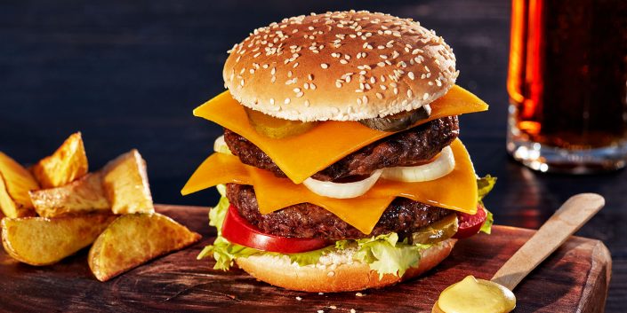 Food-Fotografie Frankfurt – Dairygold 6 | Fotografie: Hans Keller Foodaufnahme von einem Cheesburger, belegt mit Rindfleischpatty und Käsescheiben von Dirygold auf rustikalem Holzuntergrund. Daneben knusprige Potato Wedges und ein Holzlöffel mit Senf. Unscharf im Hintergrund sieht man ein Glas mit dunklem Bier.