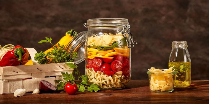 Weckglas mit gemischtem Salat auf brauner Holzplatte. Dazu verschiedene Accessoires wie ein Holzkörbchen mit Gemüse, eine Ölflasche und ein kleines Glas mit gehobelten Parmesan