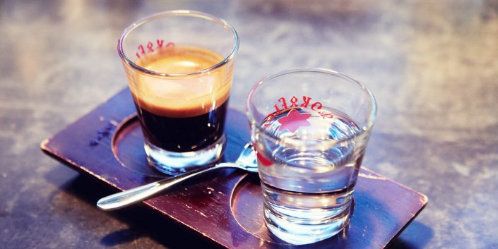 Gals mit Espresso und daneben ein Glas Wasser. Die Gläse stehen auf einem Servierbrettchen auf dem noch ein kleiner Löffel liegt.